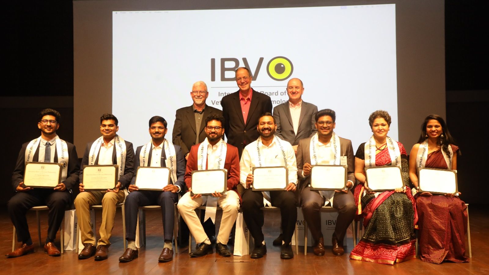 The eight new IBVO Diplomates seated on stage holding their certificates, the three founders standing behind them and the IBVO logo on the screen